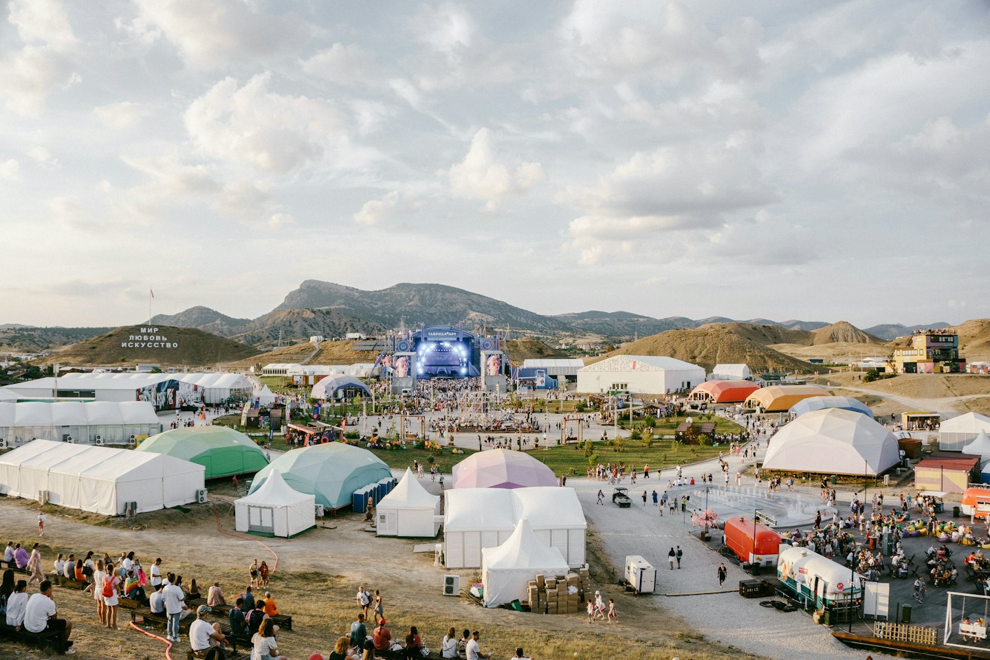 Folla di persone a un festival all'aperto con tende e montagne.